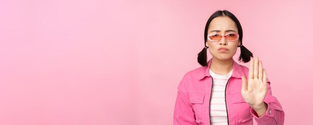 Afbeelding van een serieus, stijlvol aziatisch meisje met een zonnebril die een stop-verboden-gebaar-taboe-bord laat zien dat nee zegt over een roze achtergrond