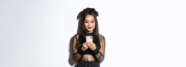 Gratis foto afbeelding van een stijlvolle aziatische vrouw in halloween-kostuum die berichten controleert op een mobiel telefoonmeisje in gotische l