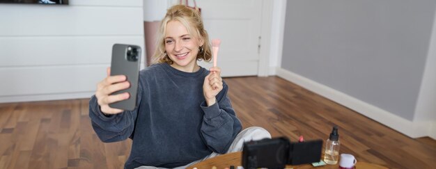 Afbeelding van een stijlvolle jonge vrouw, een social media influencer, die foto's maakt met haar mobiele telefoon terwijl ze make-up maakt.