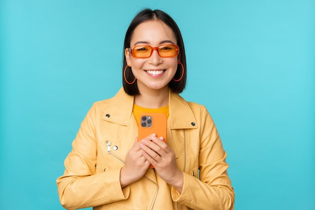 Afbeelding van een stijlvolle, mooie chinese vrouw in een zonnebril die een smartphone vasthoudt die foto's maakt op een mobiele telefoon en glimlacht die iets opneemt over een blauwe achtergrond