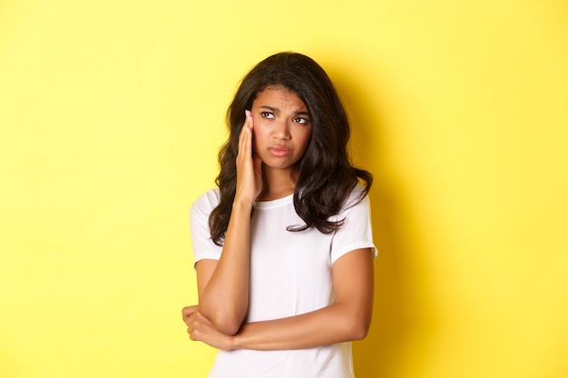 Afbeelding van een verdrietig en somber afro-amerikaans meisje, links boos kijkend en pruilend, ongemakkelijk voelend terwijl ze over een gele achtergrond staat.
