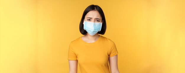 Afbeelding van een verdrietige en teleurgestelde aziatische vrouw met een medisch masker die fronst en er ontevreden uitziet tegen een gele achtergrond