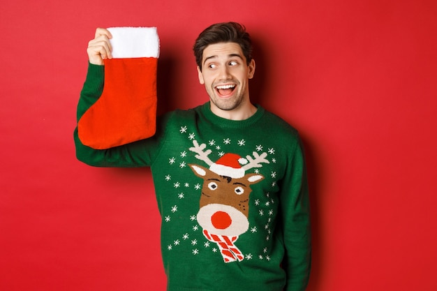 Afbeelding van een verraste en geamuseerde man in een groene trui, kijkend naar kerstsok met cadeautjes en glimlachen, staande over rode achtergrond