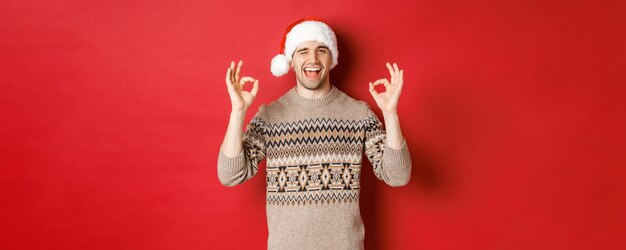 Afbeelding van een vrolijke, aantrekkelijke man in trui en kerstmuts die vrolijk kerstfeest wenst met goede tekens a