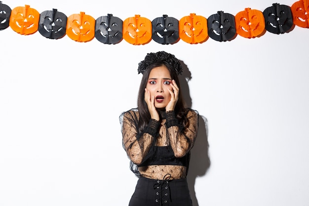 Gratis foto afbeelding van een vrouw in heksenkostuum die bang kijkt, afschuw of angst uitdrukken terwijl ze op een witte achtergrond met pompoenbanners staat, halloween vieren.