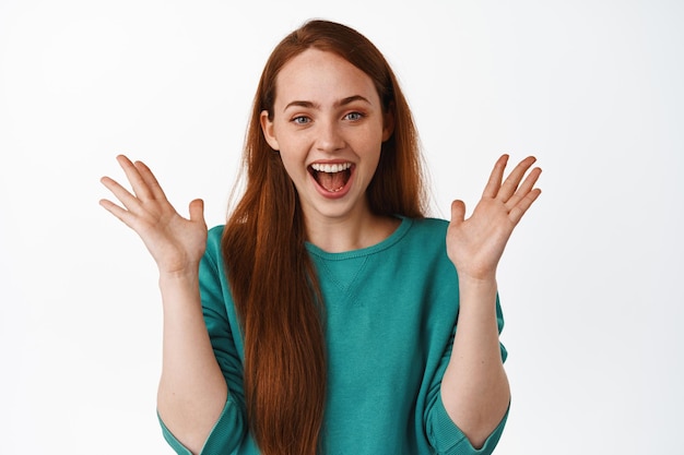 Gratis foto afbeelding van gelukkig mooi meisje met rood haar, handen klappen en vrolijk lachen, genieten van het kijken naar grappige en grappige gebeurtenis, staande op witte achtergrond