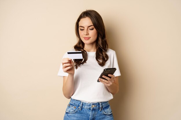 Afbeelding van jonge gelukkige vrouw die online betaalt, smartphone en creditcard vasthoudt, informatie invoert in applicatie op mobiele telefoon, staande tegen een beige achtergrond