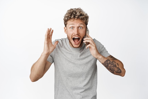 Afbeelding van jonge man praat over telefoon ontvangt een telefoontje en kijkt met ongeloof schreeuwend naar camera met opgewonden gezichtsuitdrukking witte achtergrond