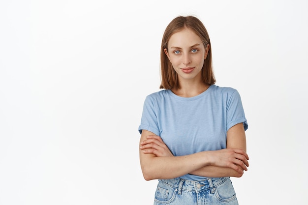 Afbeelding van jonge moderne vrouw, studente in t-shirt ziet er vastberaden en zelfverzekerd uit met gekruiste armen op de borst en brutale glimlach, staande tegen een witte achtergrond