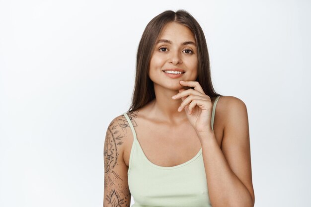 Afbeelding van jonge stijlvolle vrouw met tatoeage op arm, zomer tanktop dragen, glimlachend en haar gezicht, witte achtergrond aan te raken.