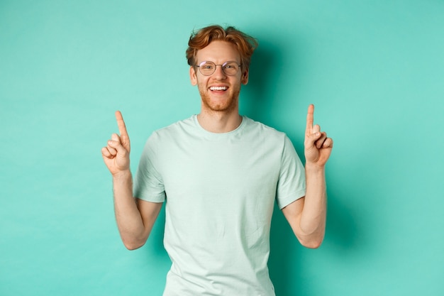 Afbeelding van knappe bebaarde man met rood haar, t-shirt en bril dragen, blij lachend en wijzende vingers omhoog, promo aanbieding tonen, staande op turkooizen achtergrond.