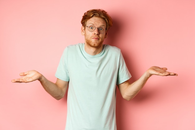 Gratis foto afbeelding van knappe roodharige man in bril en t-shirt weet niets, schouderophalend en verward wenkbrauwen optrekken, zonder idee tegen roze achtergrond.