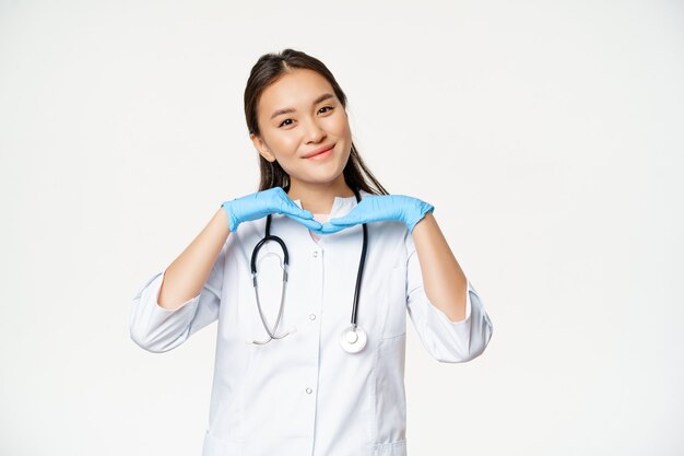 Afbeelding van lachende Aziatische arts, dermatoloog toont een gezonde huid en schattig poseren voor de camera, staande in medisch uniform op witte achtergrond.