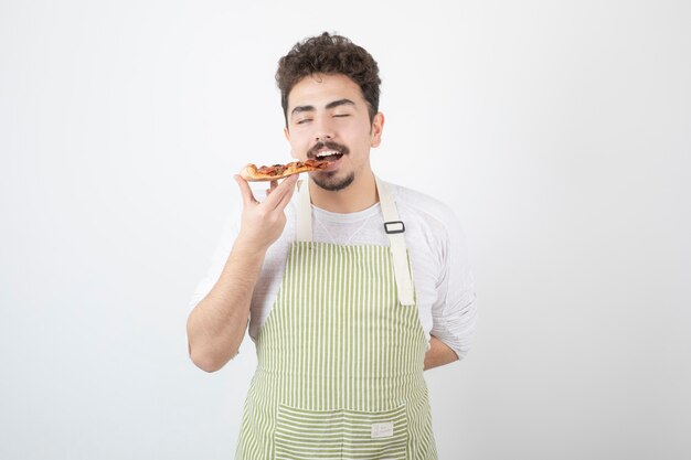 Afbeelding van mannelijke kok die een stuk pizza op wit eet