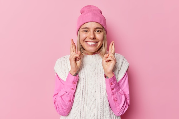 Afbeelding van mooi Europees meisje anticipeert op goed positief nieuws houdt vingers gekruist glimlacht graag draagt hoed hemd en gebreid wit vest poseert tegen roze achtergrond Mogen mijn dromen uitkomen