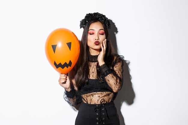 Afbeelding van mooie stijlvolle Aziatische vrouw vieren halloween, heks kostuum dragen, oranje ballon met gezicht te houden.