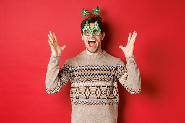 Afbeelding van opgewonden en verbaasde blanke man in feestbril, kersttrui, handen omhoog en grote aankondiging, genietend van nieuwjaarsviering, rode achtergrond.