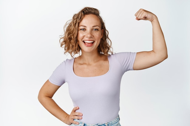 Afbeelding van positief blond meisje glimlacht, flex biceps, toont sterke armspieren, staat tegen een witte achtergrond.