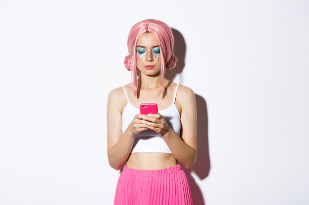 Afbeelding van stijlvolle glamour meisje in roze pruik, serieus kijken naar mobiele telefoon, permanent in party outfit.