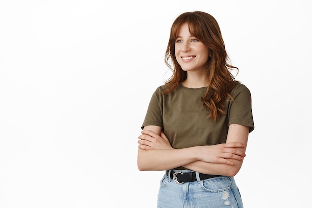 Afbeelding van stijlvolle jonge vrouw in t-shirt, armen gekruist op de borst, ontspannen en natuurlijk staan, opzij kijkend alsof ze met iemand praat, witte achtergrond.