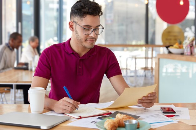 Afbeelding van tiener met trendy kapsel, draagt een optische bril, maakt records in Kladblok, houdt papier vast, gebruikt moderne technologie voor verre werk, vormt tegen café-interieur, ontwikkelt nieuwe startup