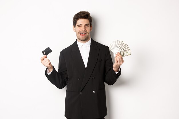 Afbeelding van zelfverzekerde zakenman in zwart pak, tevreden glimlachend en knipogend, met geld en creditcard, staande tegen een witte achtergrond.