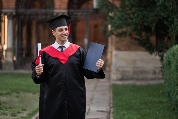 Afgestudeerde man in afstudeerjurk met diploma in zijn handen op de campus.