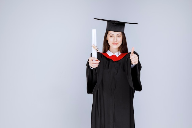 Afgestudeerde vrouwelijke student die haar diploma toont en duimen opgeeft. Hoge kwaliteit foto