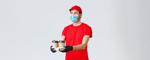 Afhaalmaaltijden en boodschappen levering covid19 contactloze bestellingen concept Aangename koerier in rood uniform gezichtsmasker en handschoenen die koffie uitdelen aan klant staan grijze achtergrond