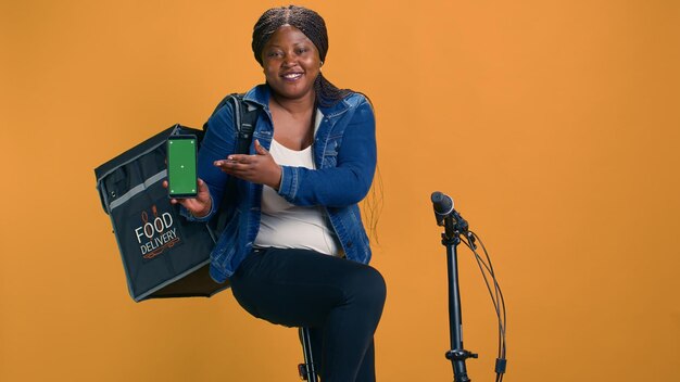 Afrikaans-Amerikaanse koerier met een mobiele telefoon met een groen scherm terwijl hij op een fiets zit Zwarte vrouw met een innovatieve bezorgapp op een mobiel apparaat met een lege mockup sjabloon