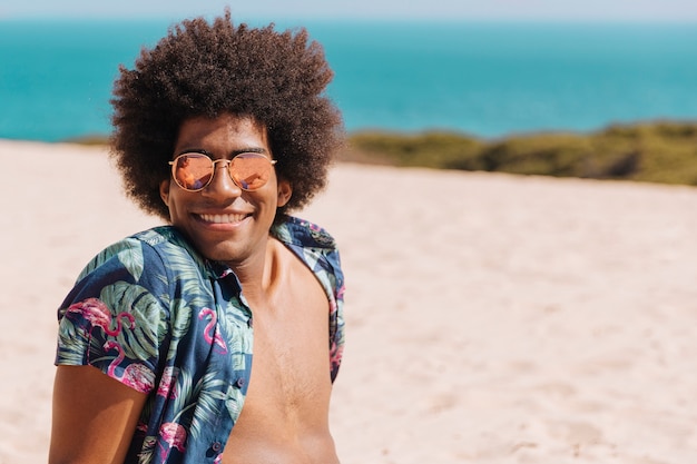Afrikaanse Amerikaanse jonge mens die in zonnebril camera op strand bekijkt