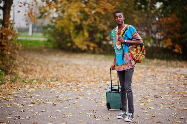 Afrikaanse man in traditioneel overhemd van Afrika op herfstpark met rugzak en koffer Emigrantenreiziger