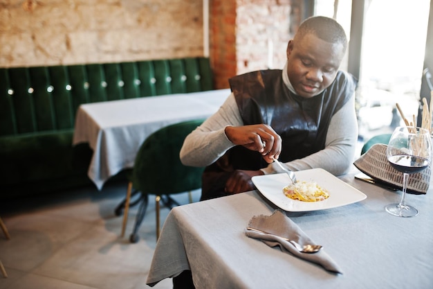 Afrikaanse man in zwarte traditionele kleding zit in restaurant en eet pasta