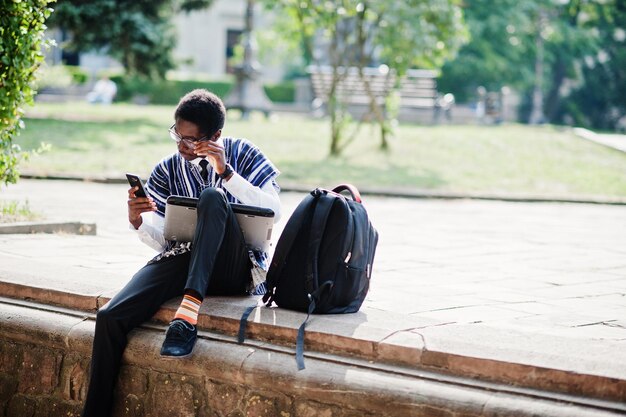 Afrikaanse man student in traditionele kleding en bril met rugzak werken op laptop met mobiele telefoon buiten