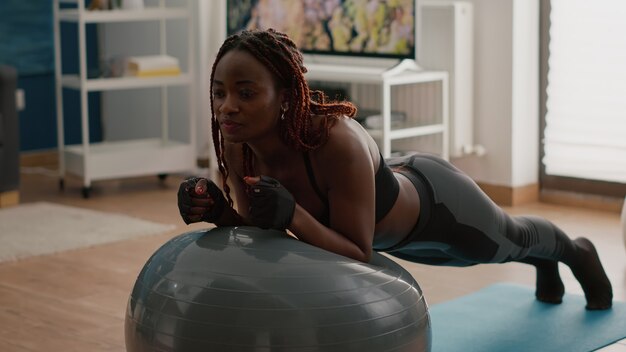 Afrikaanse slanke vrouw die buikspier uitrekt terwijl ze op yoga zwitserse bal zit en ochtendtraining doet in de woonkamer