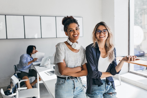 Afrikaanse vrouw in trendy casual kleding kwam naar het kantoor van vriend en poseerde met Aziatische programmeur. Charmante blonde secretaresse die lol heeft tijdens de pauze terwijl haar Japanse collega aan het werk is.