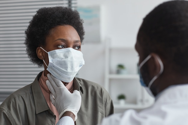 Afrikaanse vrouw met beschermend masker zit op het kantoor van de dokter terwijl de dokter haar keel onderzoekt