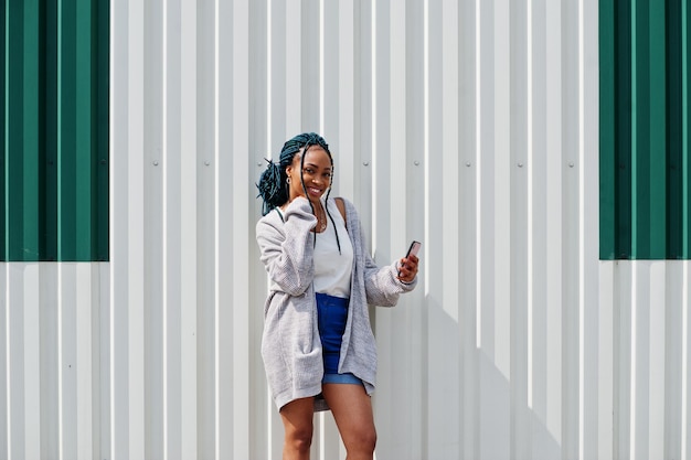 Afrikaanse vrouw met dreadlocks haar in korte spijkerbroek poseerde tegen een witte stalen muur met mobiele telefoon in de hand