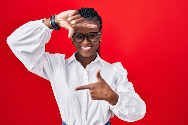 Afrikaanse vrouw met vlechten die over een rode achtergrond staan en lachend een frame maken met handen en vingers met een blij gezicht. creativiteit en fotografieconcept.