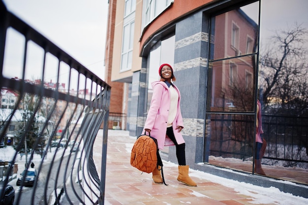 Afro-Amerikaans meisje in rode hoed en roze jas met rugzak op straat van de stad tegen voortbouwen op winterdag