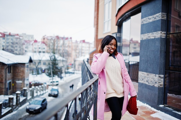 Afro-Amerikaans meisje in rode hoed en roze jas op straat van de stad tegen het voortbouwen op winterdag met mobiele telefoon