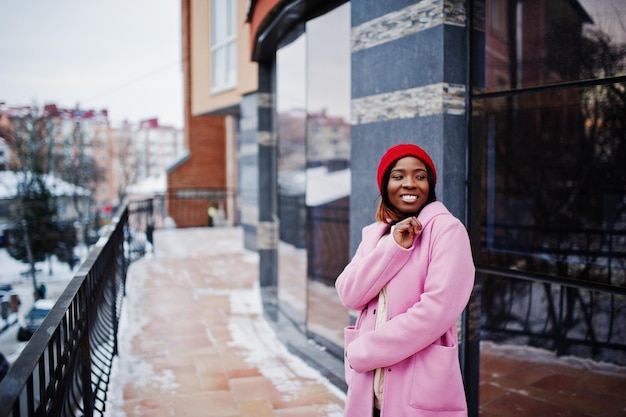 Afro-Amerikaans meisje in rode hoed en roze jas op straat van de stad tegen het voortbouwen op winterdag