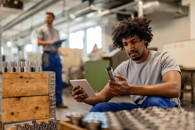 Gratis foto afro-amerikaanse arbeider die touchpad gebruikt tijdens het onderzoeken van vervaardigde roestvrijstalen staven in een fabriek