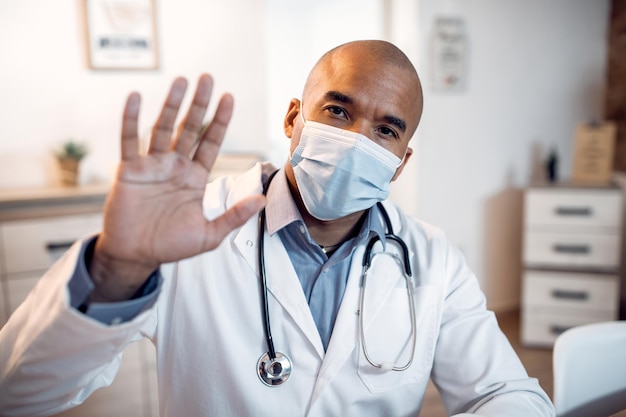 Afro-Amerikaanse arts met gezichtsmasker zwaaiend tijdens telefonische vergadering vanuit de kliniek