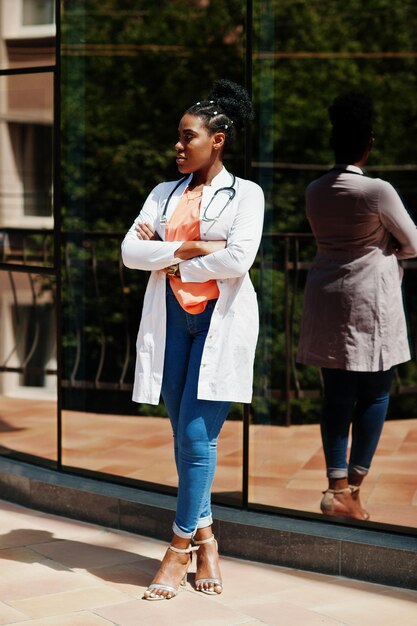 Afro-Amerikaanse arts-vrouw bij laboratoriumjas met stethoscoop buiten tegen kliniek