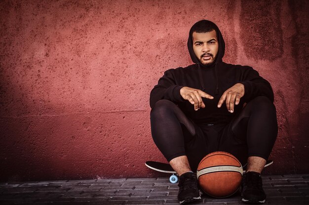 Afro-Amerikaanse bebaarde man gekleed in een zwarte hoodie en sportbroek zittend op een skateboard met basketbal en leunend op de muur.