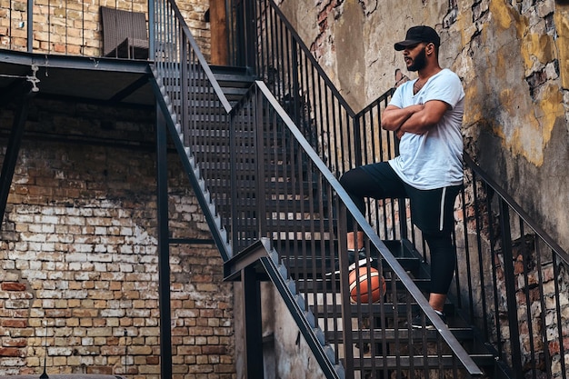 Afro-Amerikaanse bebaarde streetball-speler met pet gekleed in sportkleding houdt een basketbal vast die op de trap staat en houdt een been op een bal in een getto.