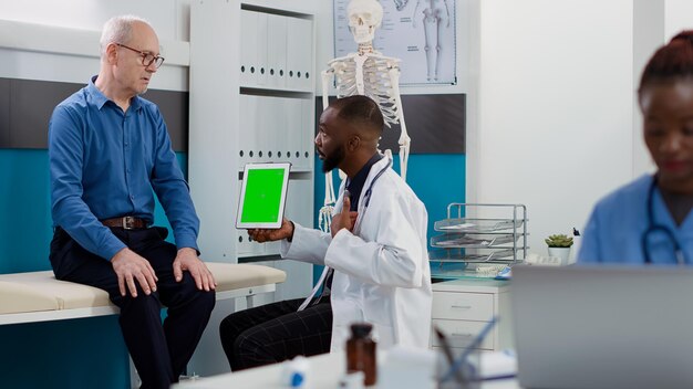 Afro-Amerikaanse dokter wijzend op tablet met groen scherm, kijkend naar chromakey-sjabloon met oude patiënt. Arts en man met behulp van geïsoleerde mockup achtergrond en lege copyspace.