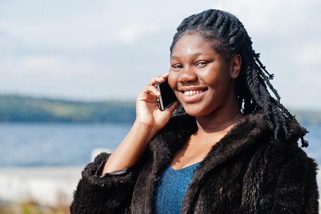 Afro-Amerikaanse donkere huid plus size model poseerde in een blauwe glanzende jurk en zwarte bontjas tegen de zeekant spreken op de telefoon