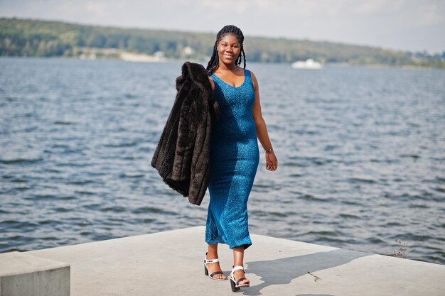Afro-Amerikaanse donkere huid plus size model poseerde in een blauwe glanzende jurk en zwarte bontjas tegen de zeekant